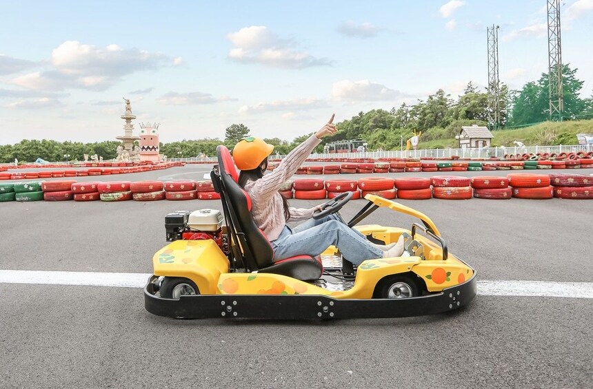 A person in a yellow go-kart on a track with red barriers, a statue, and a tower in the background. 4