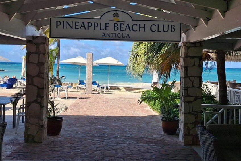 A beach club entrance with a sign, outdoor seating, umbrellas, and a view of the beach and ocean. 2
