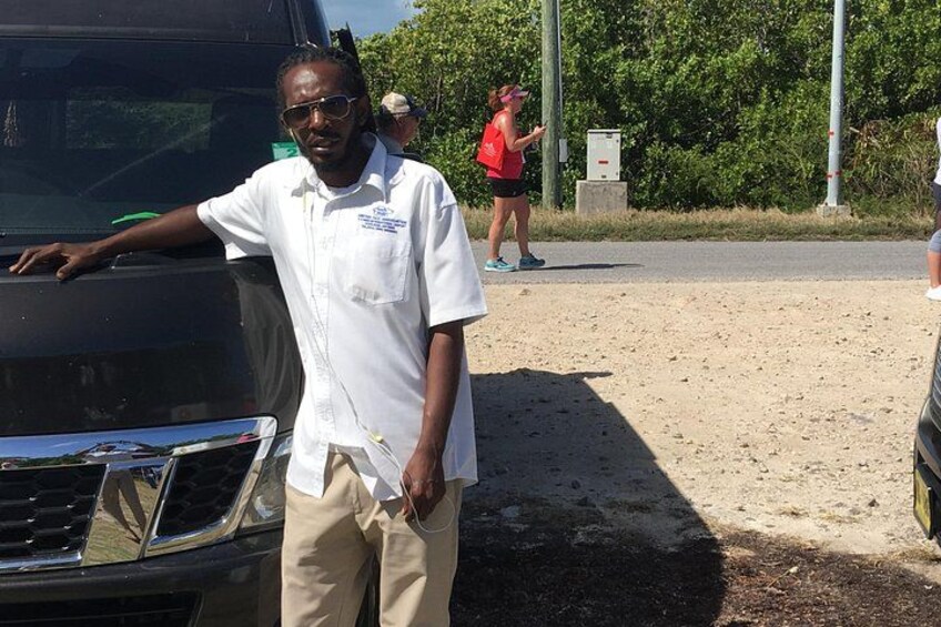 A person standing next to a black vehicle, a dirt road, and a green area with trees. 5