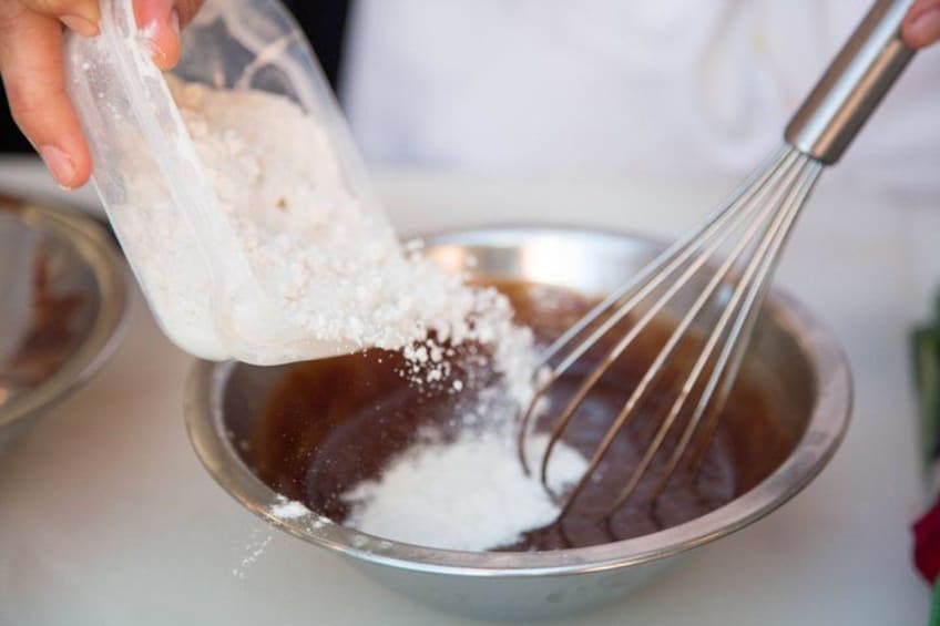 A person pouring a white powder into a metal bowl with a whisk. 3