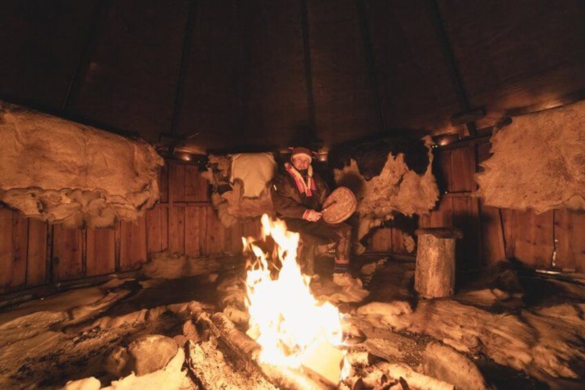 A traditional wooden interior with a central fire, stone walls, and a wooden bench. 1