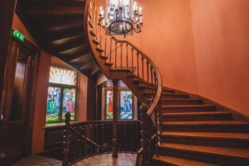 A grand staircase with ornate railings and a chandelier, leading to a room with stained glass windows. 5