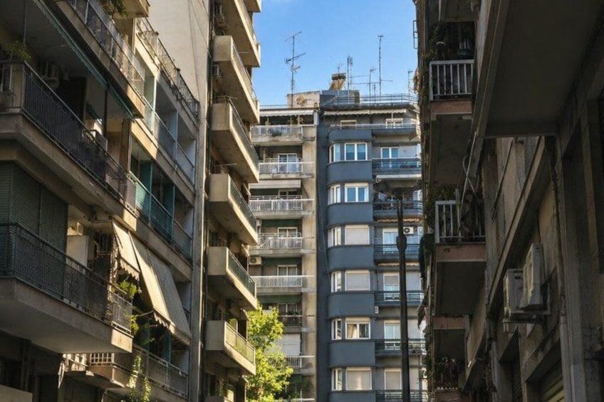 A multi-story residential building with balconies, a narrow street, and a clear blue sky. 3