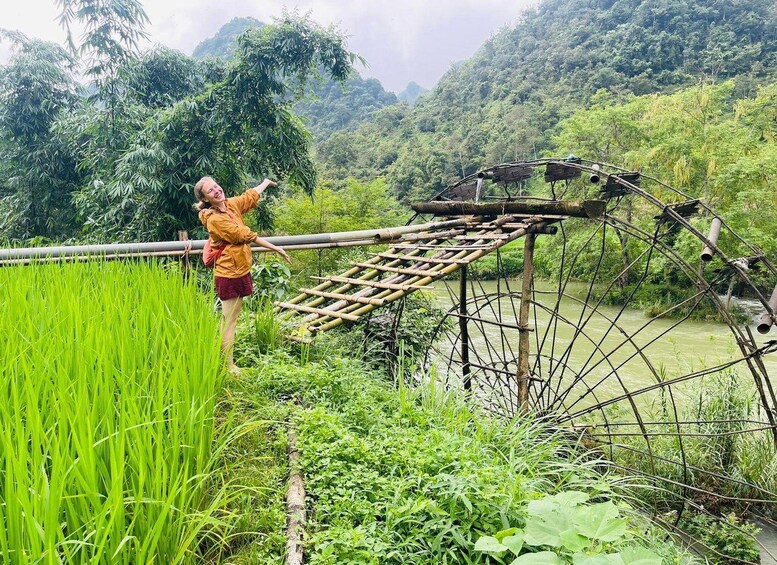 Eine Person steht auf einer Bambusbrücke über einem Fluss, umgeben von üppiger Vegetation und einer gebirgigen Landschaft. 2