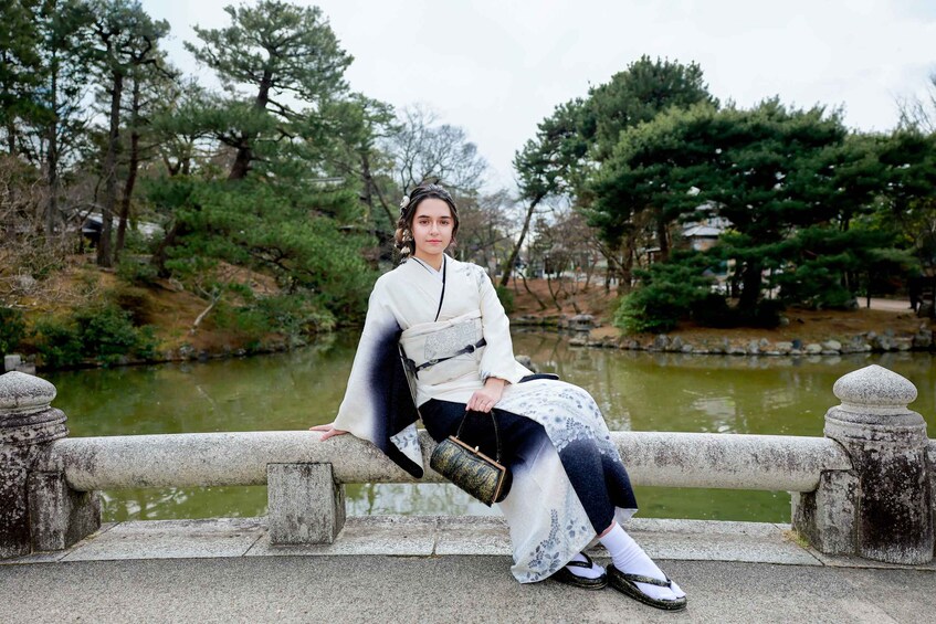 A person in traditional attire sitting on a stone bridge with a pond and trees in the background. 2