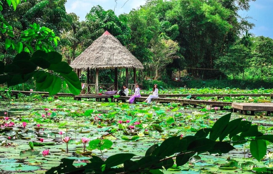 A thatched-roof pavilion surrounded by a pond with lotus flowers, with people sitting on a wooden platform. 4