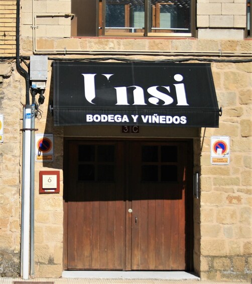 A storefront with a black awning displaying the name 'Unsi' and the words 'Bodega y Vinedos' underneath. There are two signs on either side of the door indicating parking restrictions. 2