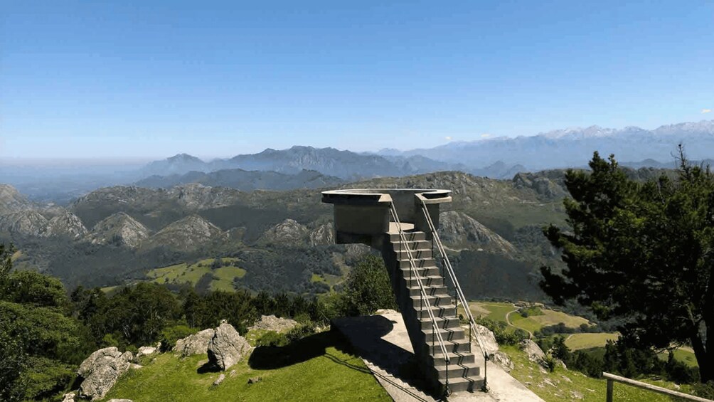 A concrete viewing platform with stairs leading up to it, overlooking a mountainous landscape with green valleys and distant snow-capped peaks. 2