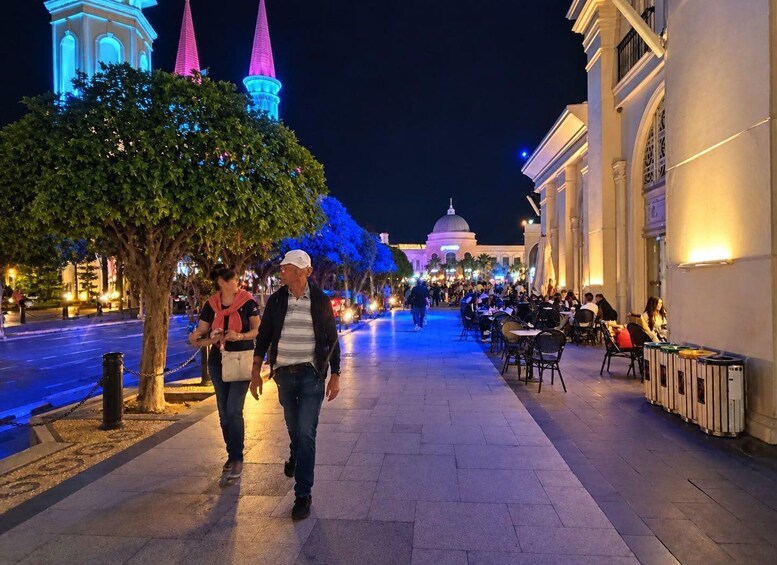 A nighttime city street with illuminated buildings, trees, and outdoor seating at a restaurant. 2