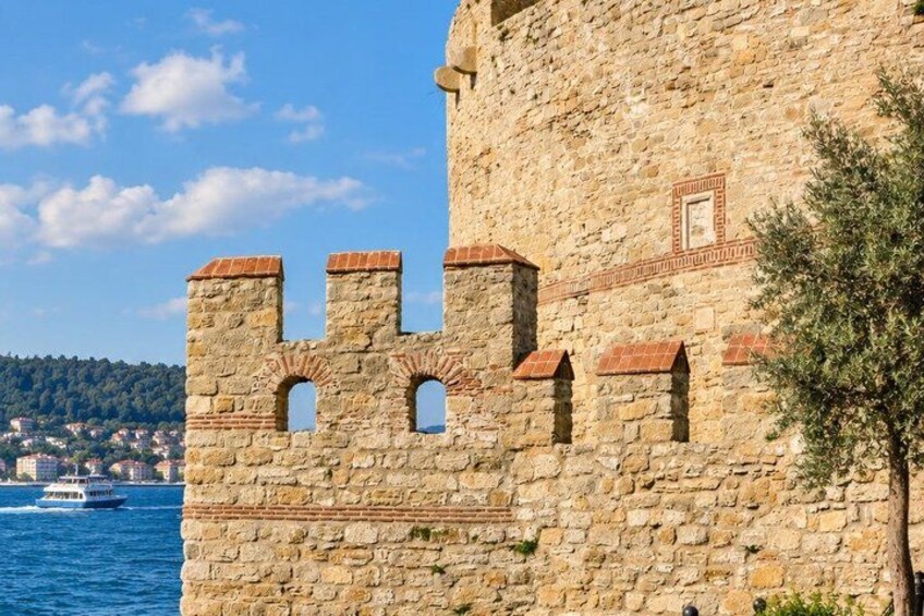 A stone castle wall with crenellations, a clear blue sky, and a body of water with a boat. 2