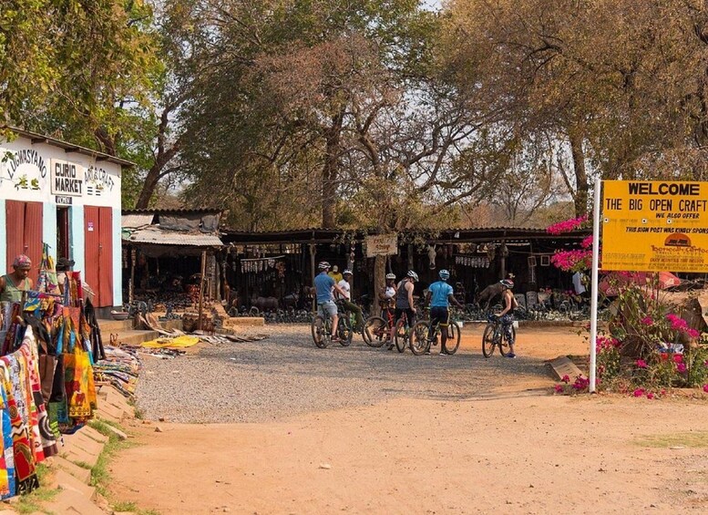 A group of cyclists on a dirt road, a market stall with colorful fabrics, and a welcome sign for The Big Open Craft. 3