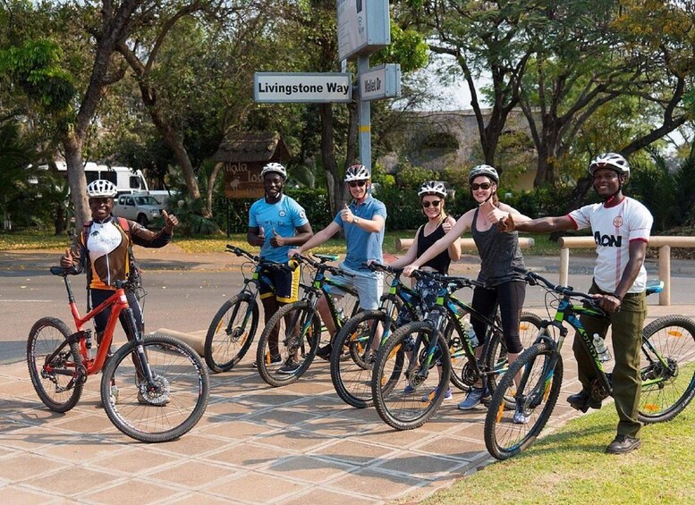 A group of cyclists on a paved path with trees and a sign reading 'Livingstone Way' in the background. 2
