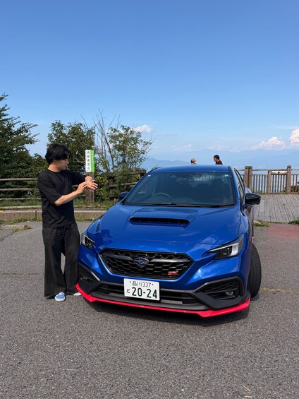 A blue car parked on a road with a person standing beside it, a signpost in the background, and a view of the sky and distant mountains. 2