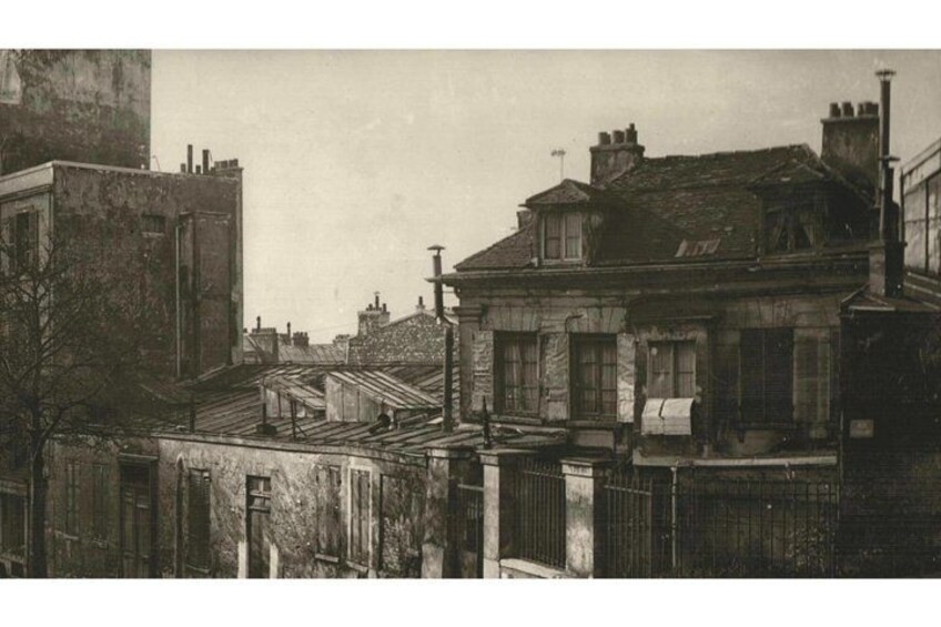 A historic urban street with old buildings, including a prominent two-story house with a pitched roof and a chimney. 2