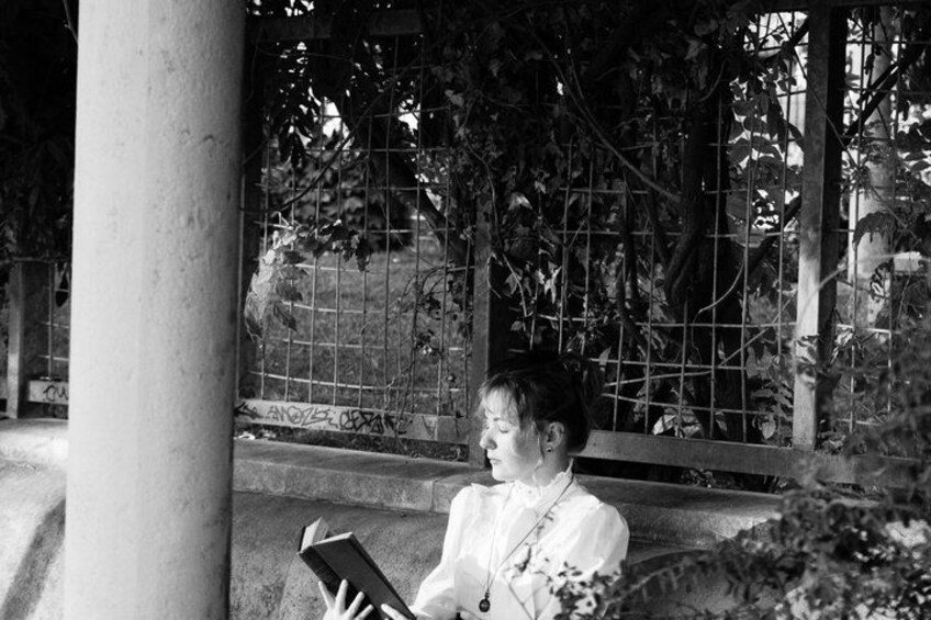 A person sitting on a bench reading a book in a shaded area with a metal fence and trees in the background. 4
