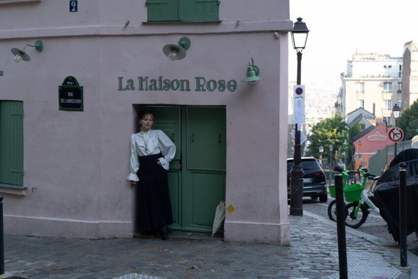 A building named 'La Maison Rose' with a green door and shutters, a person standing in front, and a street with parked cars and a green bicycle. 5