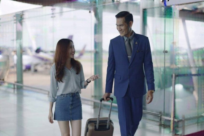 A man in a blue suit with a suitcase, a woman in a denim skirt and blouse, and an airport interior with glass partitions and airplanes in the background. 1