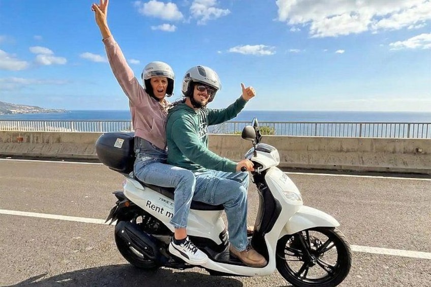 Two people on a white scooter with 'Rent me' written on it, riding on a road with a coastal view. 1