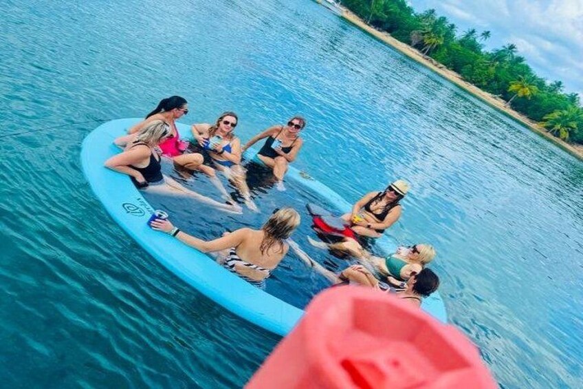 A group of people on a blue inflatable raft in a body of water surrounded by greenery. 3