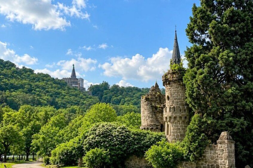 A castle with towers, lush green trees, and a distant church on a hill. 2