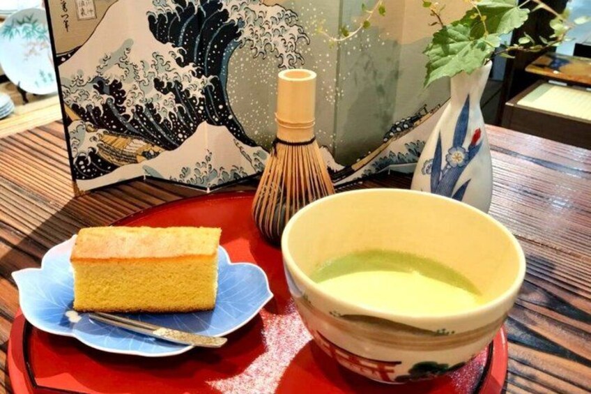 A piece of cake on a blue plate, a bowl of green tea, and a traditional Japanese whisk on a wooden table. 4