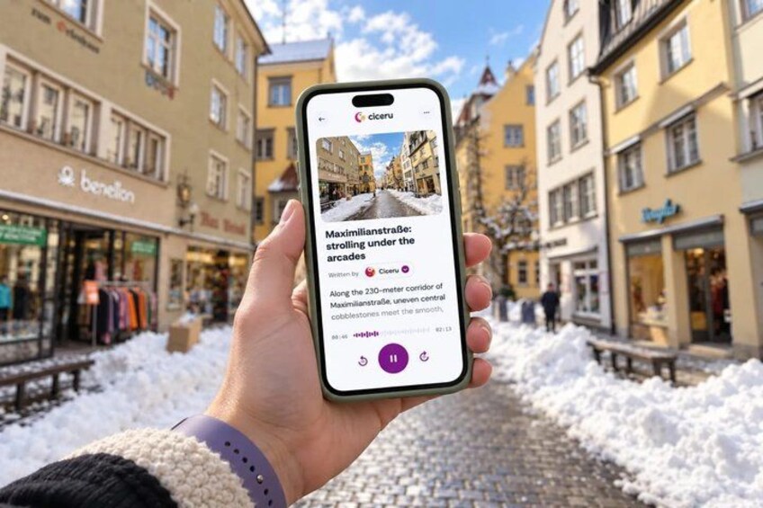 A hand holding a smartphone displaying a street view of Maximilianstrasse with snow on the ground. 1