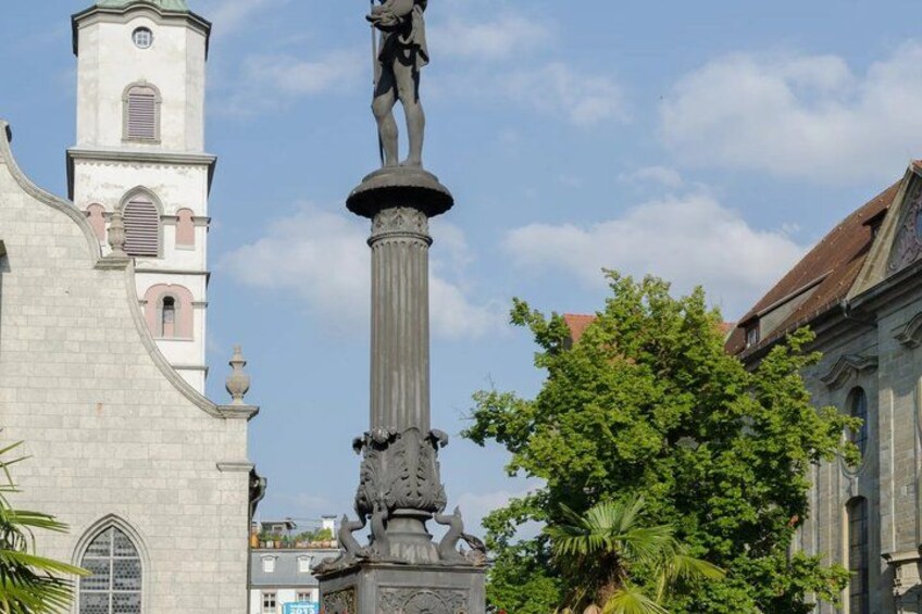 A historical building with a clock tower, a statue on a pedestal, and lush green trees. 2