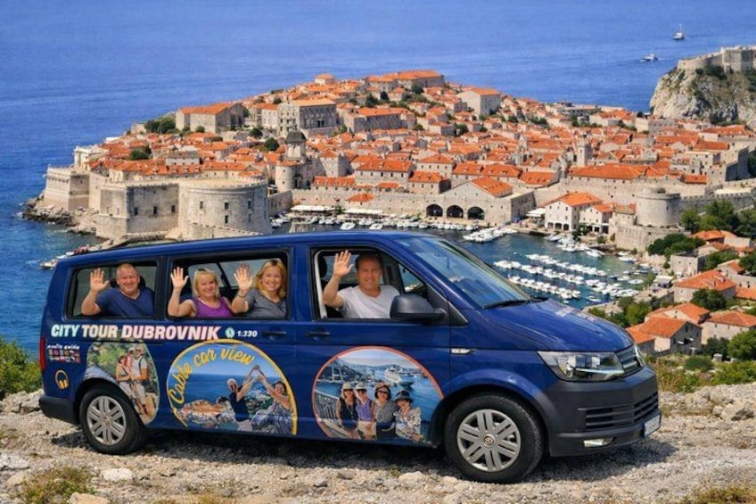 A blue van with tourists waving, parked on a rocky terrain with a coastal cityscape in the background. 1