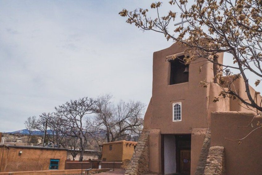 A traditional adobe building with a bell tower, surrounded by bare trees and a mountainous backdrop. 4