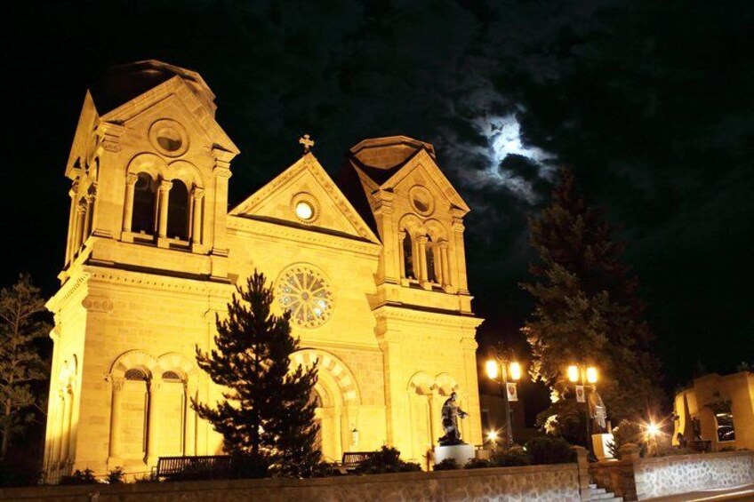 A lit-up church with a clock, a statue, and a moon in the sky. 1