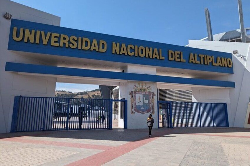 The entrance to Universidad Nacional del Altiplano with a blue and white gate, a person walking, and a clear sky. 5
