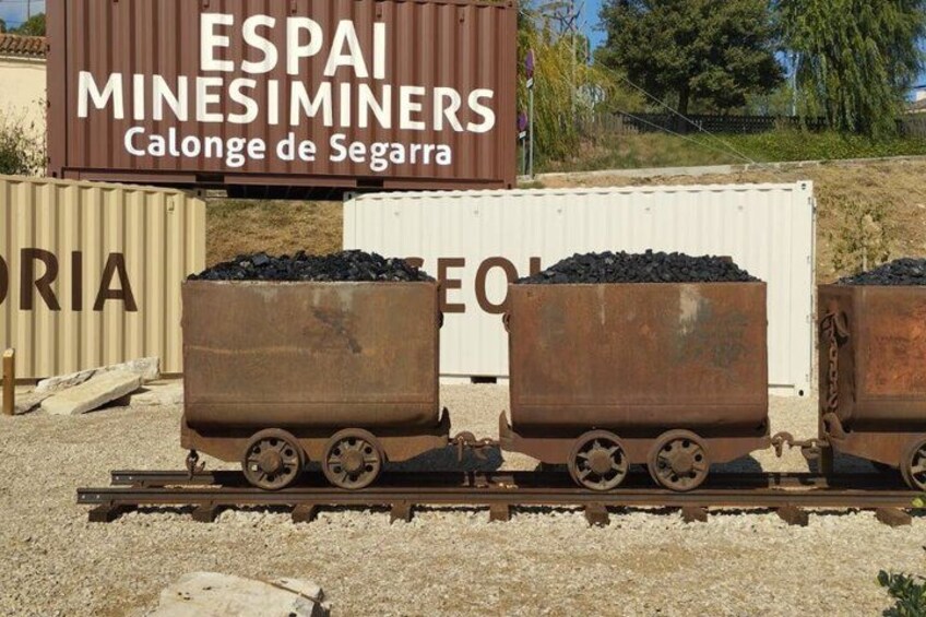 A mining site with a sign reading 'ESPAI MINESIMINERS Calonge de Segarra' and two rusty railway carts filled with coal. 2