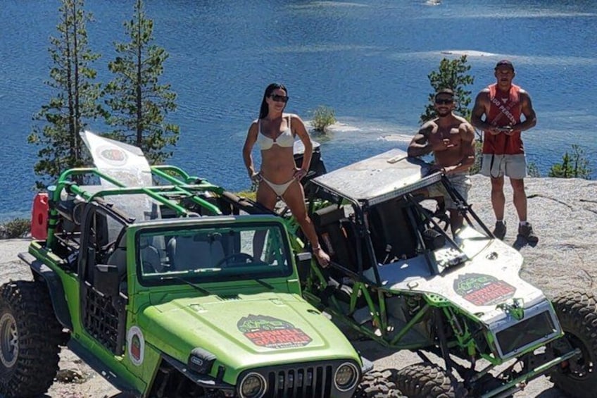 A green off-road vehicle with a surfboard, a woman in a bikini, and two men standing on a rocky shore by a body of water. 5