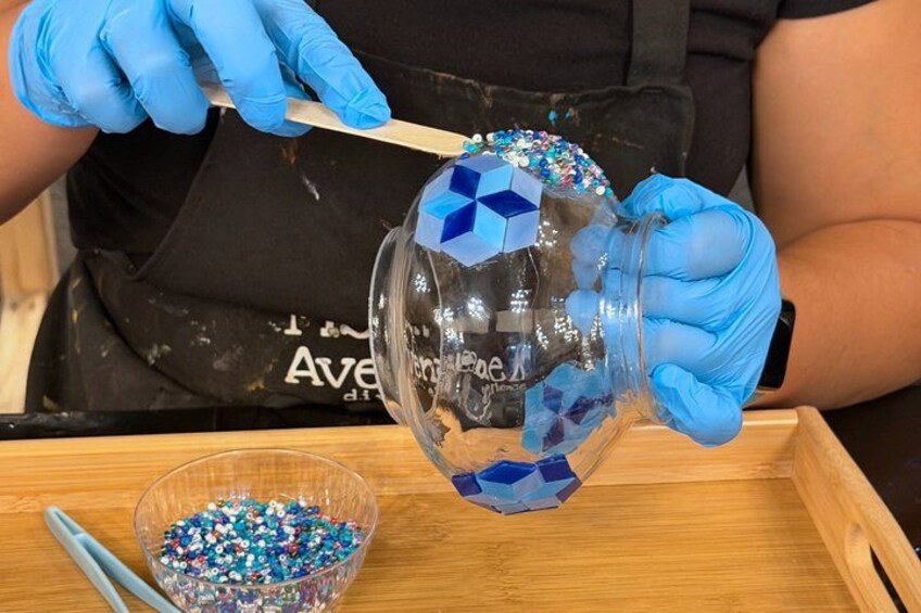 A person wearing a black apron and blue gloves, holding a glass bowl with blue and white beads. 4