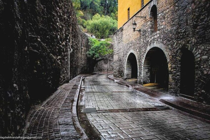 A narrow cobblestone street flanked by stone walls and arched doorways, leading through a hilly landscape. 5