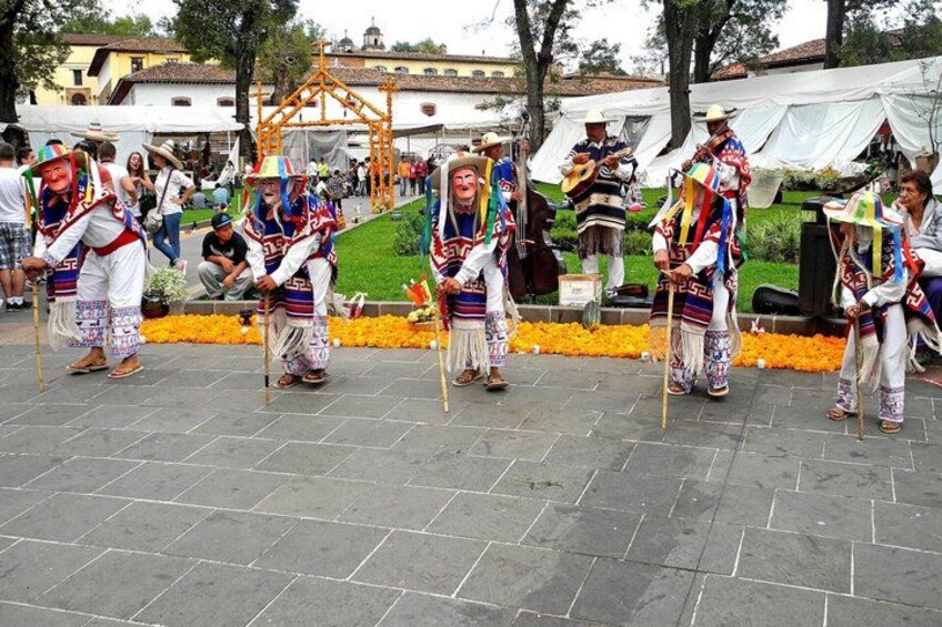 A group of performers in traditional attire dancing in an outdoor setting with a stone-paved area and a backdrop of buildings and trees. 2