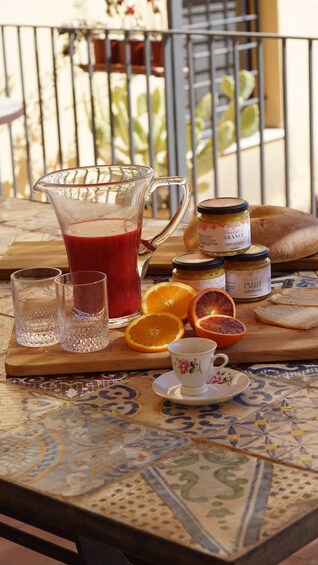 Een tafel met een karaf rode sap, twee glazen, een pot marmelade, oranjeslices, een kop en brood. 4