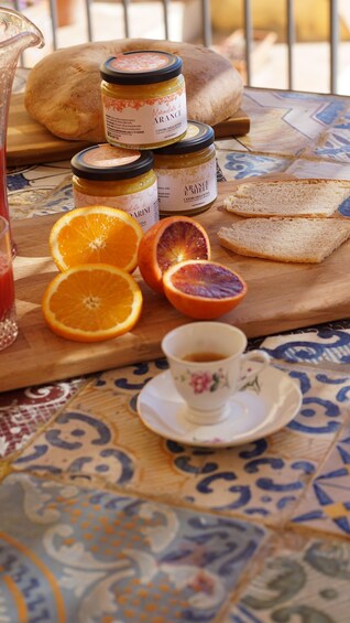 Een tafel met potten marmelade, schijfjes sinaasappel, een kop thee en brood. 3