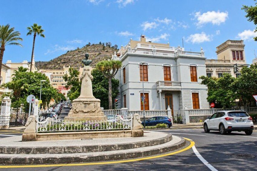 A roundabout with a statue, a white building with red shutters, palm trees, and a hilly background. 4