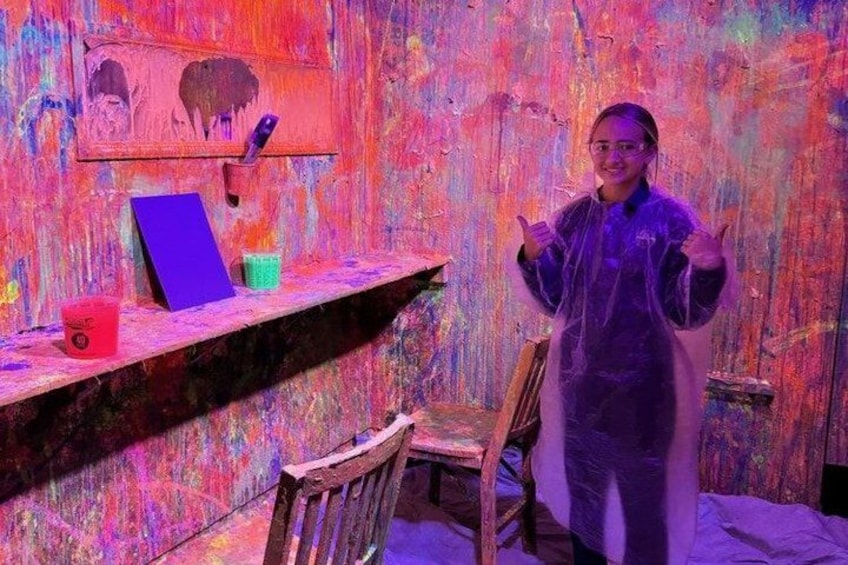 A person in a protective suit standing in a room with a colorful, textured wall and a wooden chair. 3