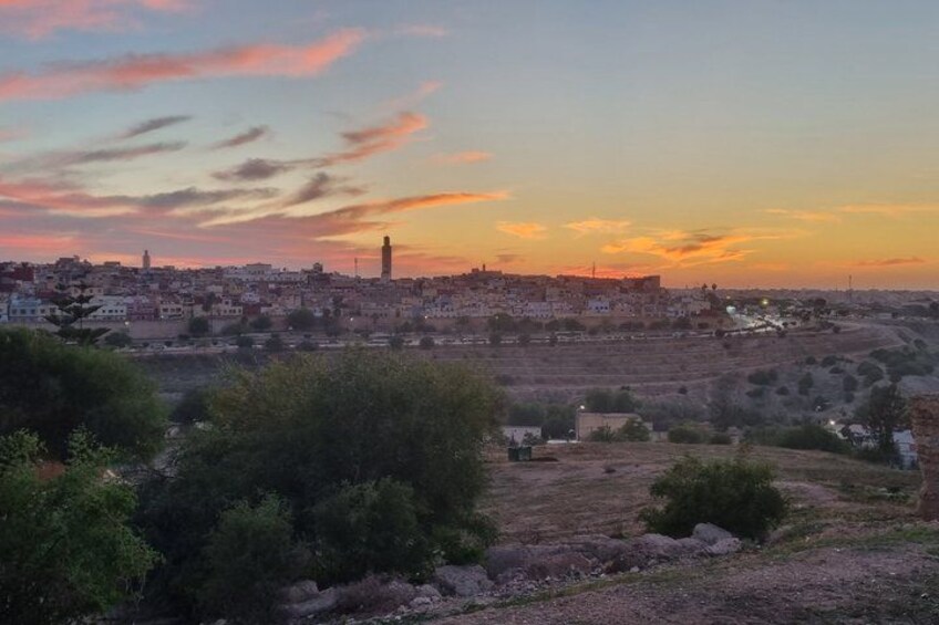 A cityscape at sunset with a prominent tower, rolling hills, and sparse vegetation. 2