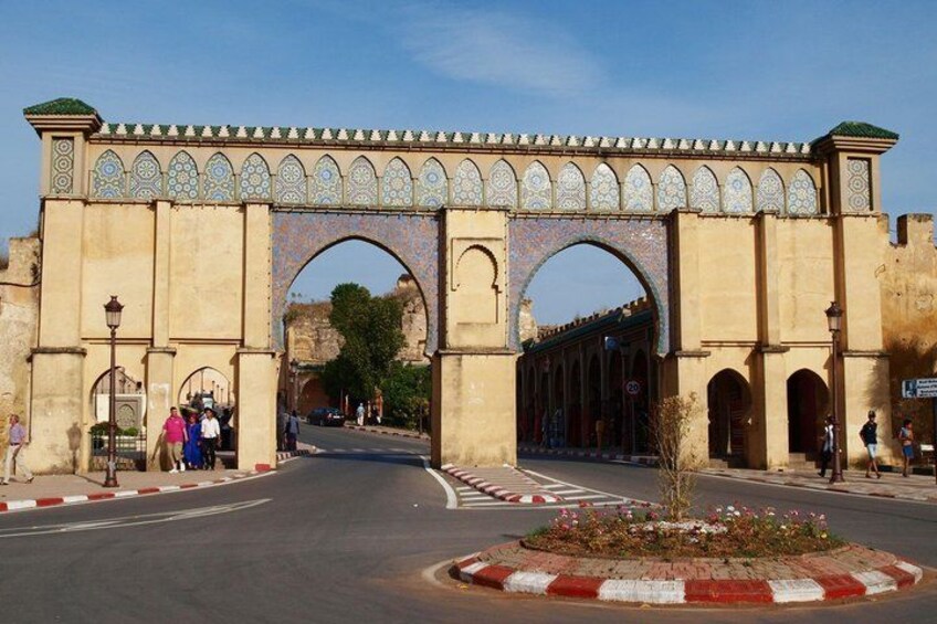 A historic archway with intricate tile work, a paved road, and a small flower bed. 4