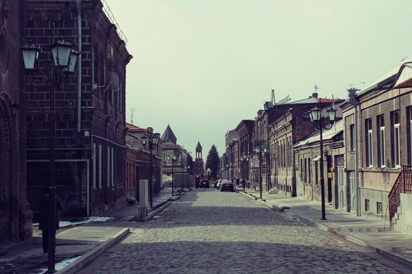 A cobblestone street lined with historic buildings, a church spire in the distance, and a parked car. 4