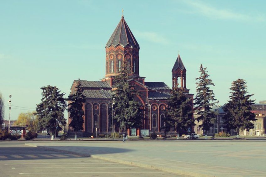 A large church with a tall bell tower, surrounded by trees and a wide street. 1