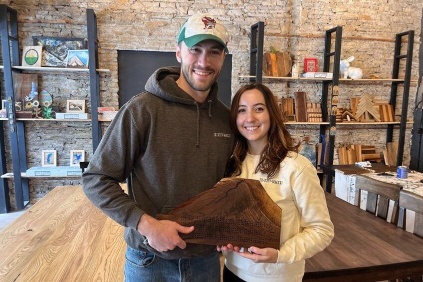 Two people holding a wooden object in a workshop with shelves of wood and tools. 1