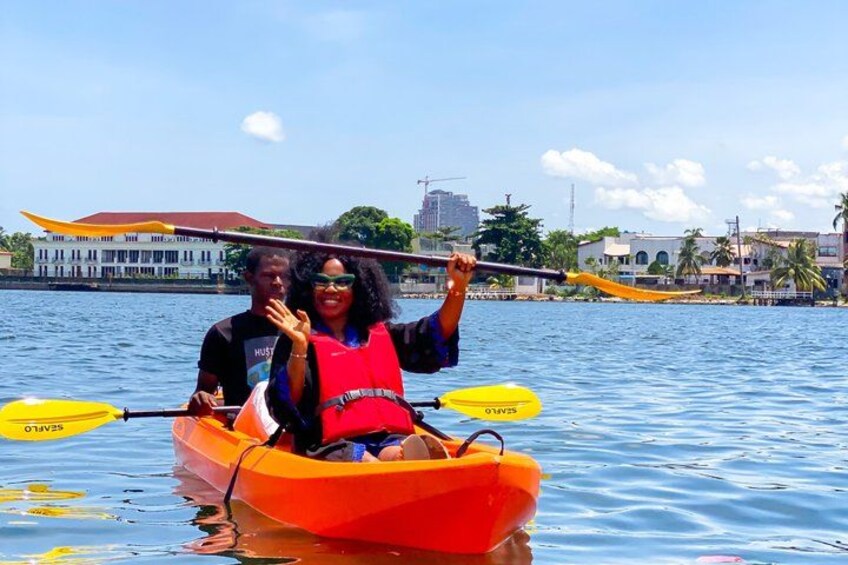 Two people kayaking on a calm body of water with buildings and palm trees in the background. 2