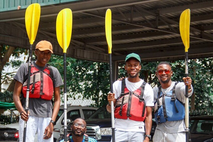 Four people wearing life jackets and holding paddles, standing under a covered area with trees in the background. 5