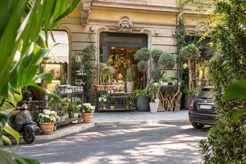 A street with a shop selling plants, a parked car, and a scooter. 3