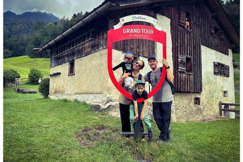A family posing in front of a rustic building with a sign reading 'Grand Tour of Switzerland.' 1