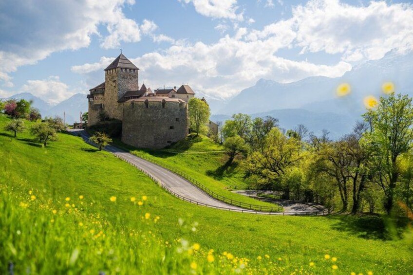 A castle on a hill with a winding road leading to it, surrounded by green fields and mountains in the background. 3